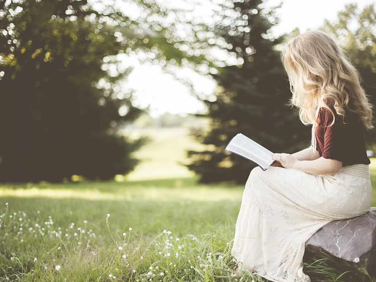 Image of a woman sitting on a rock in a field reading her bible